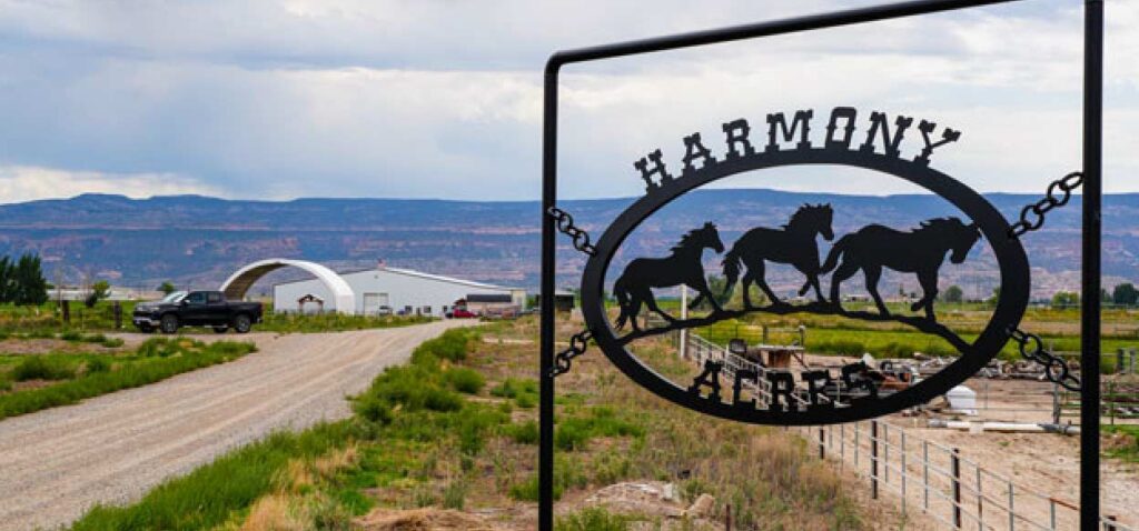 Sign reading "Harmony Ranch" with horse silhouettes in the foreground, gravel road leading to a building and mountain landscape in the background.